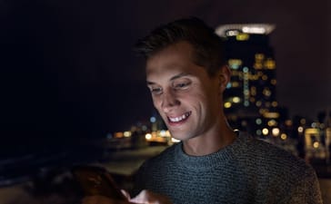 Man using a smartphone at night in a city setting with blurred lights in the background.
