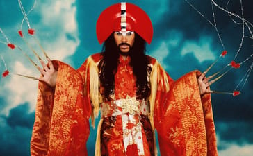 Empire Of The Sun promotional portrait of Luke Steele as the Emperor in a red headdress and orange robes against a blue background.