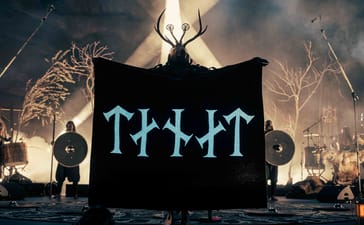 An antlered Heilung performer holds a black banner onstage, with shield-bearing performers behind them.