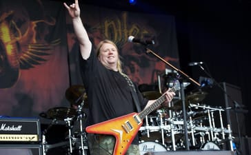 Karl Sanders smiles and raises the horns while holding an orange electric guitar onstage.