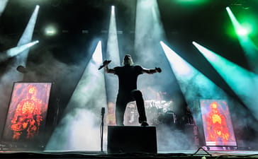 Meshuggah vocalist Jens Kidman holds a microphone with his arms outstretched beneath white and green stage lights.
