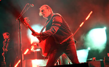 Josh Homme leans forward while playing guitar under red stage lights.