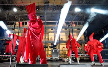 TISM performing in red costumes under bright stage lights at the Sidney Myer Music Bowl.