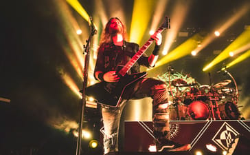Reece Scruggs plays guitar with one foot raised beneath yellow stage lights.