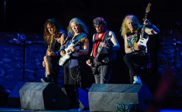Four members of Iron Maiden playing bass and guitars onstage under blue lighting.
