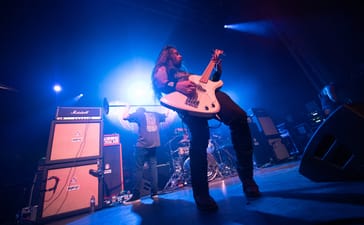 Power Trip bassist Chris Whetzel performing under blue stage lighting, with Riley Gale in the background.