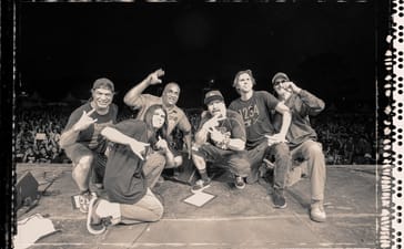 Black-and-white group photograph of Infectious Grooves posing on stage, with a crowd behind them.