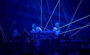 Underworld’s Rick Smith and Karl Hyde performing behind electronic equipment beneath blue lighting and white laser beams.