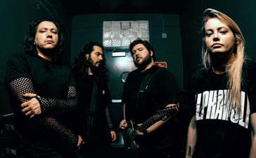 • • A dark promotional portrait of the four members of the metal band VCTMS standing in a dimly lit backstage room or hallway, wearing all-black clothing.