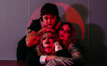 Three members of Vipersnatch pose together under red lighting against a grey wall.