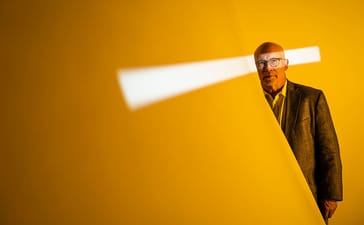 Portrait of Alex Gibney against an orange background crossed by a strip of white light.