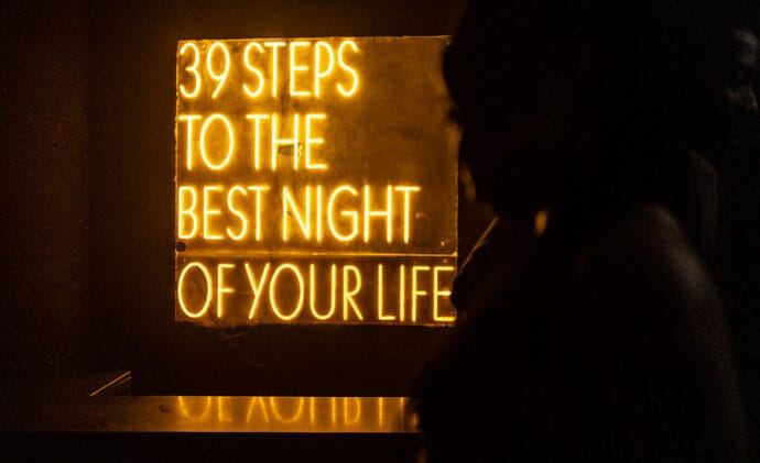 Yellow neon sign reading ‘39 steps to the best night of your life,’ glowing in a dark room with a silhouetted figure.
