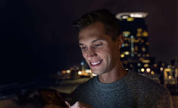 Man using a smartphone at night in a city setting with blurred lights in the background.