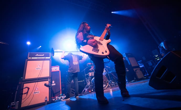 Power Trip bassist Chris Whetzel performing under blue stage lighting, with Riley Gale in the background.