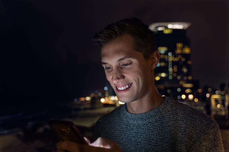 Man using a smartphone at night in a city setting with blurred lights in the background.