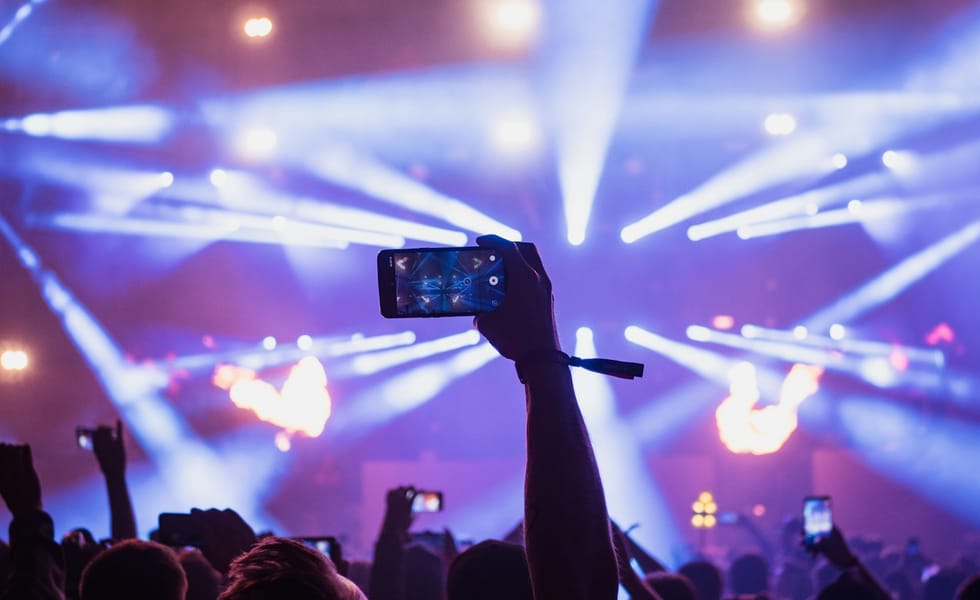 Audience member filming a live concert on a smartphone beneath purple stage lights.