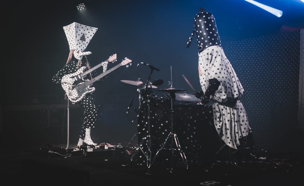 Angine de Poitrine performing in polka-dot costumes and oversized masks, with a double-neck guitar and drum kit.