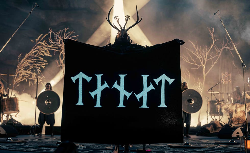 An antlered Heilung performer holds a black banner onstage, with shield-bearing performers behind them.