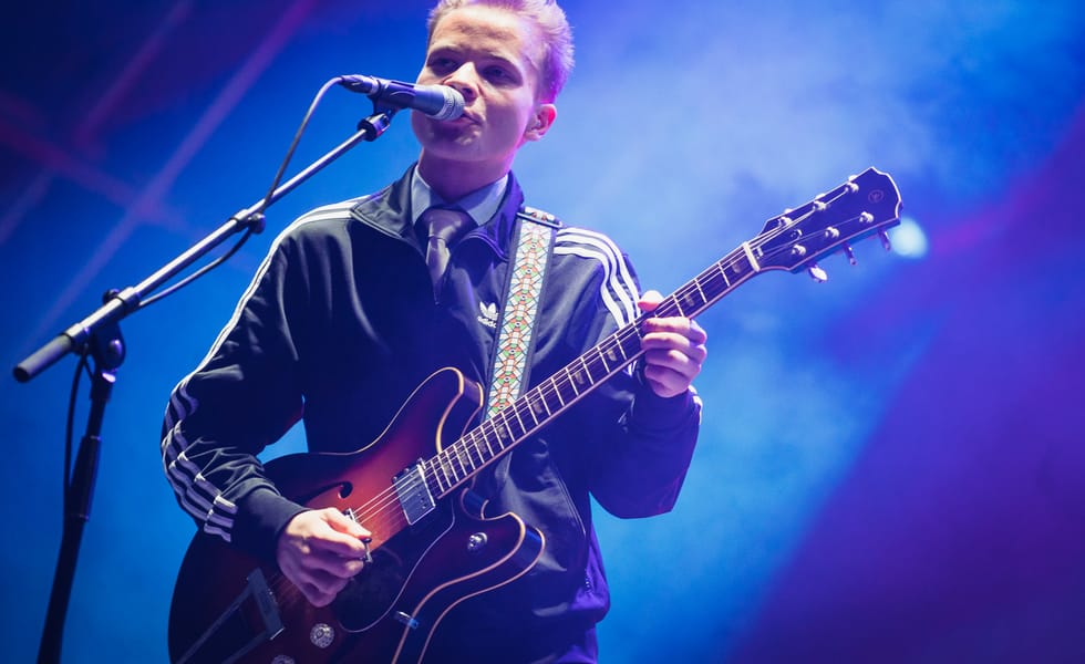 Geordie Greep singing into a microphone and playing guitar under blue stage lighting.