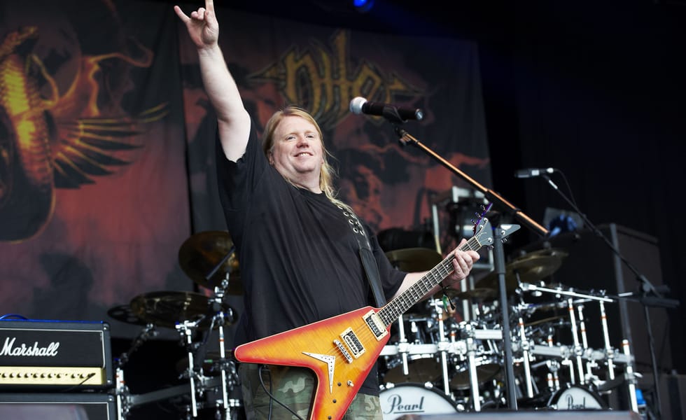 Karl Sanders smiles and raises the horns while holding an orange electric guitar onstage.