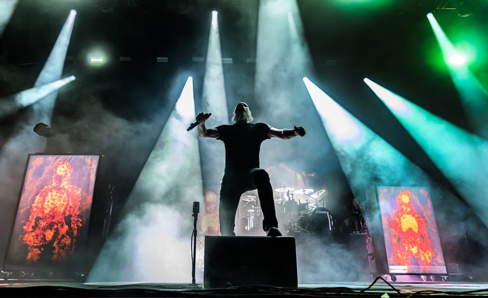Meshuggah vocalist Jens Kidman holds a microphone with his arms outstretched beneath white and green stage lights.