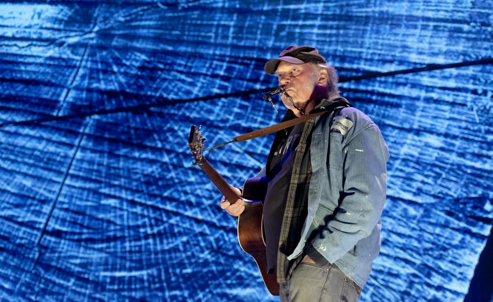 Neil Young holding an acoustic guitar and wearing a harmonica holder against a blue stage backdrop.