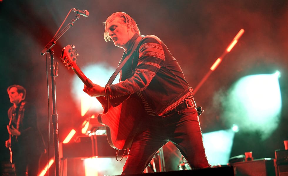Josh Homme leans forward while playing guitar under red stage lights.
