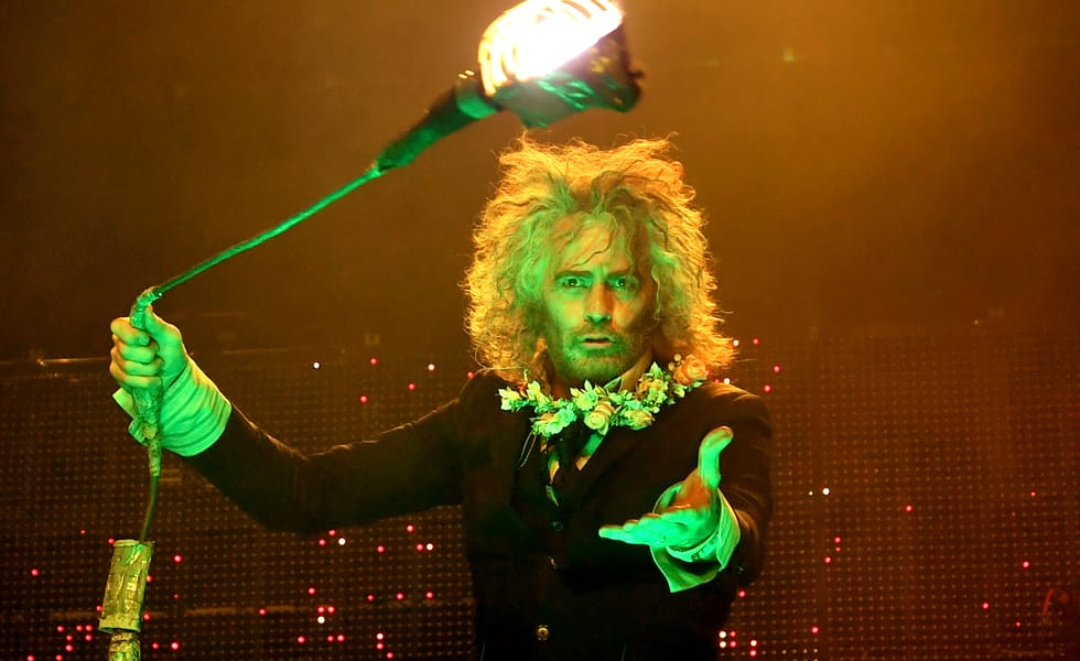 Wayne Coyne of The Flaming Lips performs under green and orange stage lighting, holding a theatrical prop towards the audience.