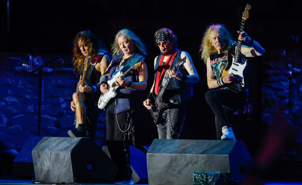 Four members of Iron Maiden playing bass and guitars onstage under blue lighting.