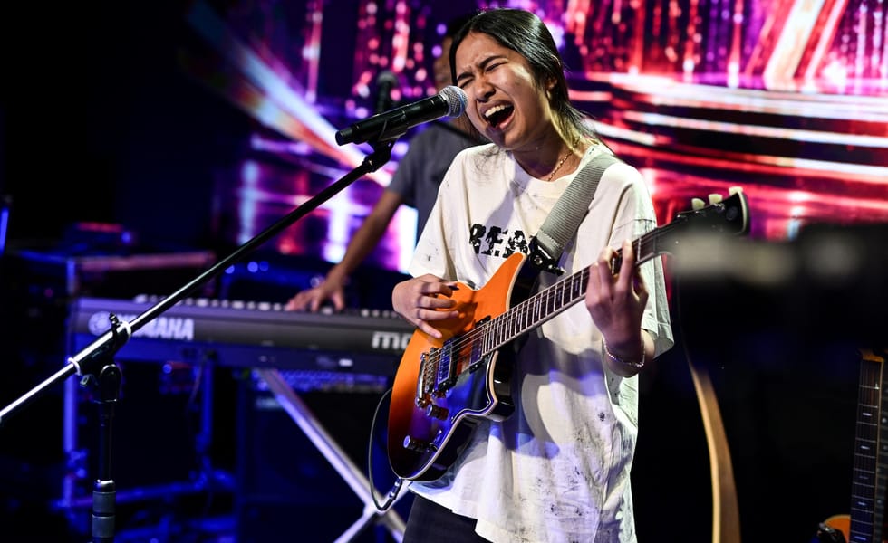 Nene Royal performs onstage, singing into a microphone while playing an orange electric guitar.