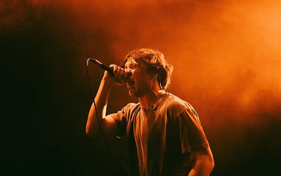 One Step Closer vocalist Ryan Savitski singing into a microphone under orange stage lighting.