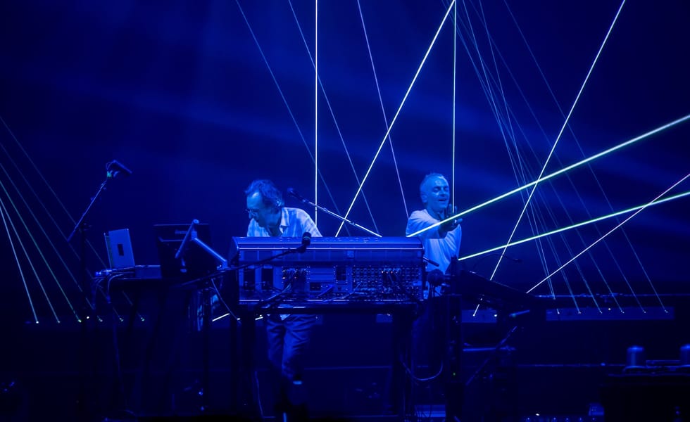 Underworld’s Rick Smith and Karl Hyde performing behind electronic equipment beneath blue lighting and white laser beams.