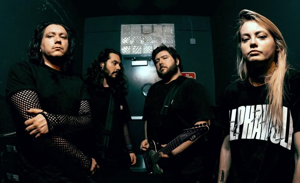 • • A dark promotional portrait of the four members of the metal band VCTMS standing in a dimly lit backstage room or hallway, wearing all-black clothing.