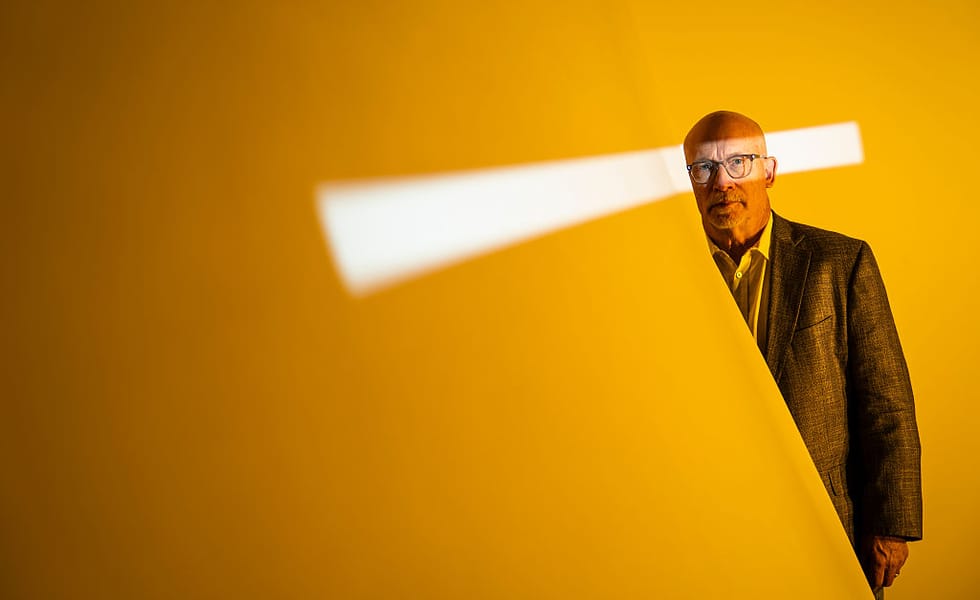 Portrait of Alex Gibney against an orange background crossed by a strip of white light.